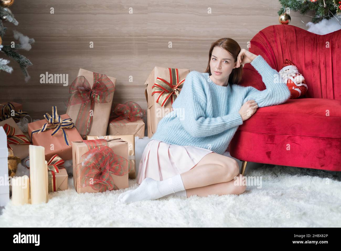 Ritratto di Capodanno della bella giovane donna caucasica in maglione azzurro, gonna bianca e calzini. Adorabile ragazza seduta vicino a scatole regalo con archi. Foto Stock