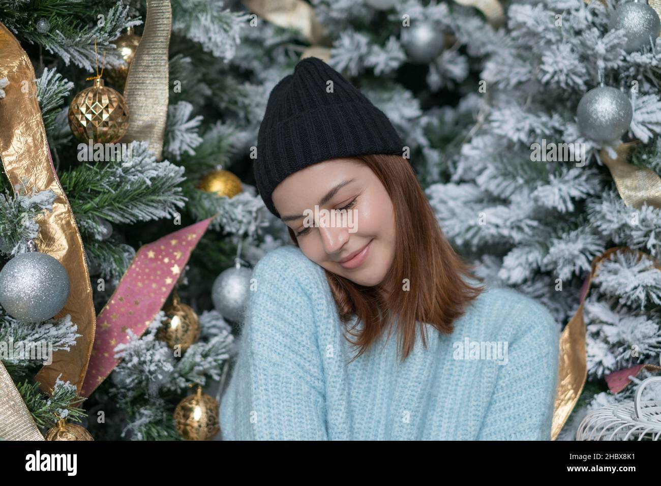Ritratto di Capodanno della bella giovane donna caucasica in morbido maglione azzurro e cappello. Adorabile sorridente ragazza seduta vicino decorato albero di Natale Foto Stock