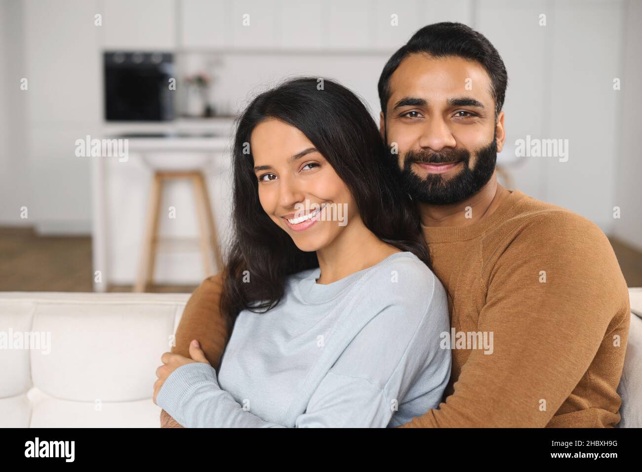 Primo piano di felice coppia interrazziale in posa mentre si siede al divano con sfondo cucina. Felici i proprietari di un nuovo appartamento sorridente e abbracciato davanti alla fotocamera Foto Stock