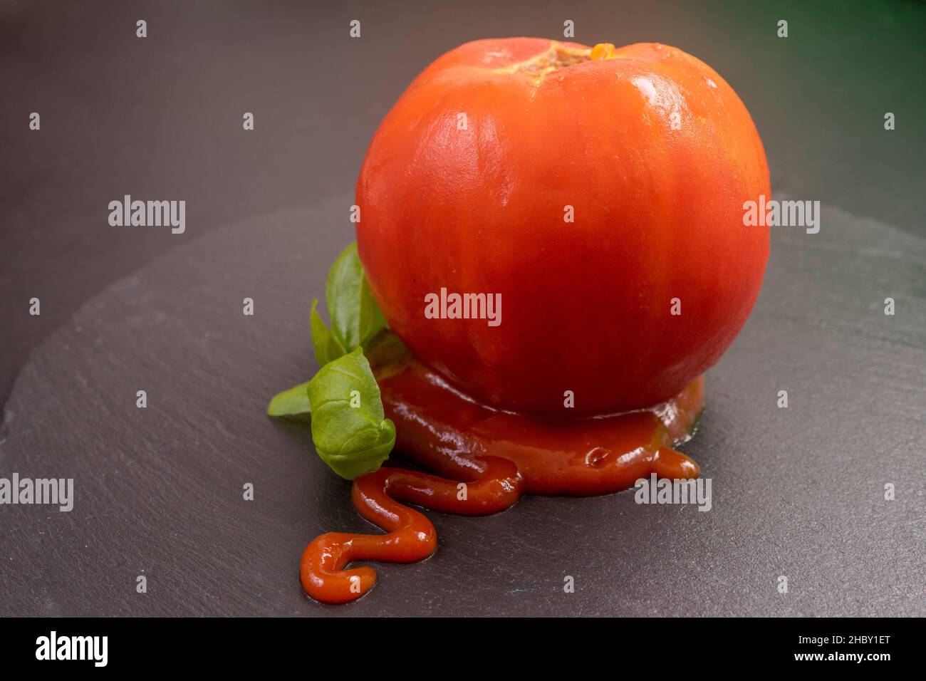 pomodoro pelato con ketchup e foglie di basilico, isolato su una piastrella di pietra nera Foto Stock
