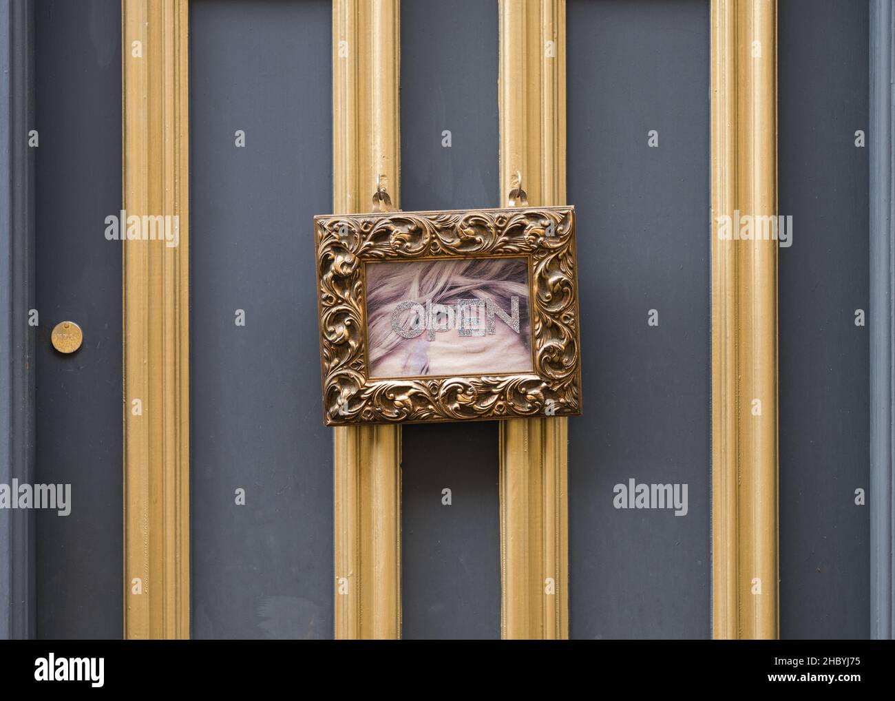 Cartello aperto con cornice in oro appeso alla porta d'epoca retrò. Cartello del negozio aperto per lavoro. Porta grigia e telaio ornato in foglie d'oro. Foto Stock
