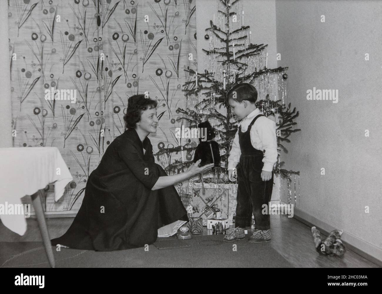 Natale 1962 in Germania. Una giovane, graziosa madre nel suo vestito festivo presenta al suo figlio non di fronte all'albero di Natale. Foto Stock