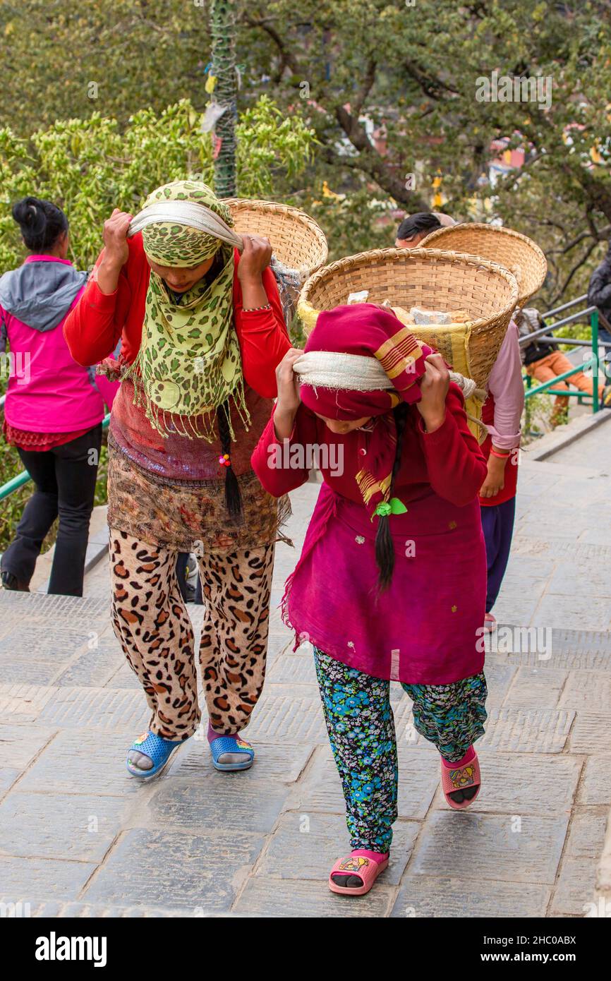 Due giovani donne nepalesi trasportano pesanti cesti di pietre sulle loro spalle su scale usando una tumpline a Kathmandu, Nepal Foto Stock