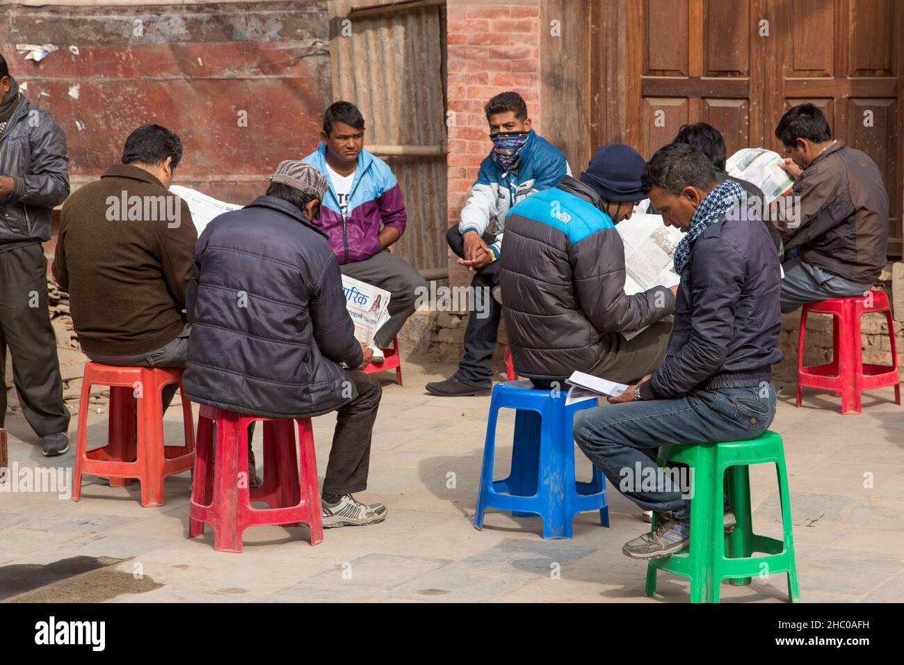 Un gruppo di uomini nepali in abito occidentale seduta su sgabelli e lettura giornali in una piazza a Kathmandu, Nepal. Foto Stock