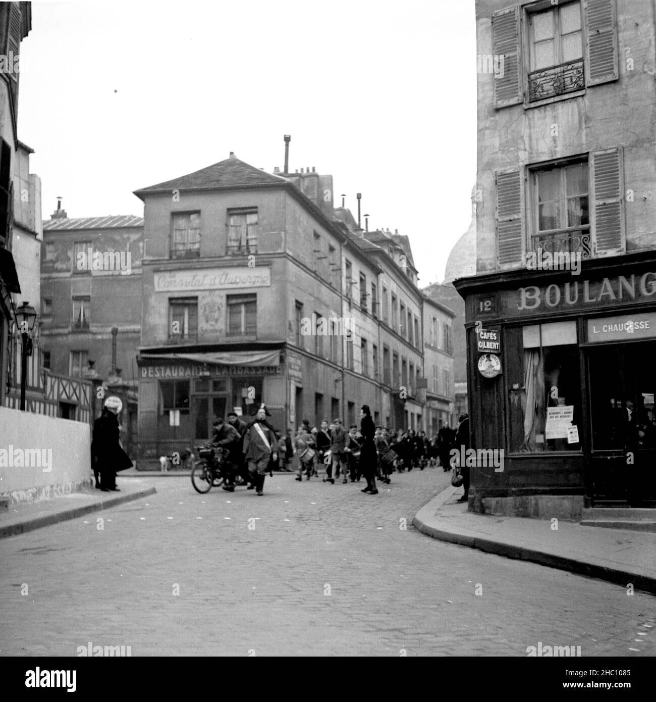 Una processione nel 1945 a Parigi Montmartre proviene dalla direzione di Sacre-Cœur camminando lungo Rue Norvins e girando a sinistra in Place Jean-Baptiste Clement. Guidare è un uomo anziano che indossa un cappello Napoleone, un mantello, e stivali da equitazione. Sigaro in bocca, una spada pende sul suo lato sinistro. Sembra aver perso il braccio sinistro. Dietro di lui in marcia c'è un gruppo di ragazzi che battono i tamburi. Una Marina degli Stati Uniti ha arruolato l'uomo orologi da un lato e Una donna orologi e poi si unisce alla parte anteriore del gruppo. La sagoma della cupola del Sacre-Coeur è in vista, così come il Consulat d’Auvergne e il Ristorante l’Ambassade. Foto Stock