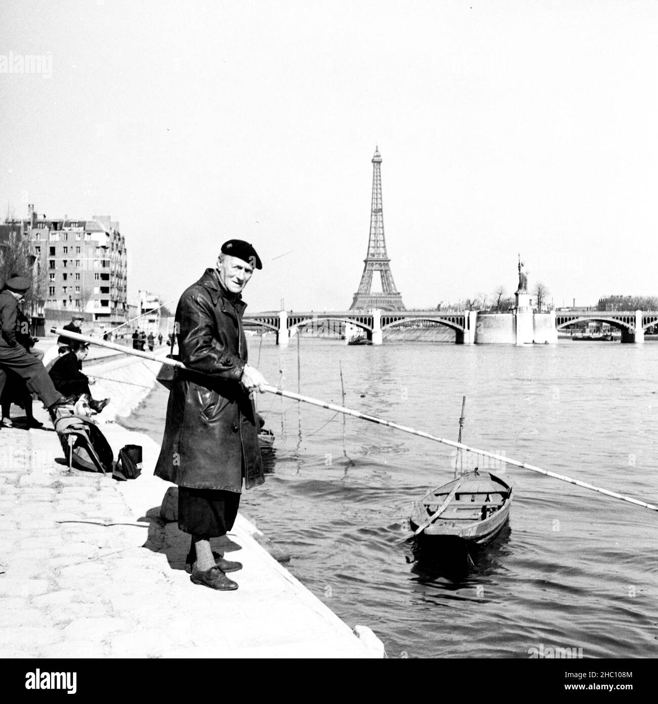 Parigi pescatori sulla Senna guardando la macchina fotografica - Pont Mirabeau, marzo 1945. Il suo vestito tradizionale include un cappotto di pelle, berretto e pantaloni a forma di culotte sopra la sox di lana. Un cannello da pesca è appeso alla spalla. La sua canna da pesca è molto più lunga di quanto sia alto. Pali vicino sul fiume sono barche a remi e altri pescatori. La Statua della libertà Petite è di fronte alla macchina fotografica da Pont de Grenelle. Alle spalle si vedono il Pont de Bir Hakeim e la Torre Eiffel. Foto Stock