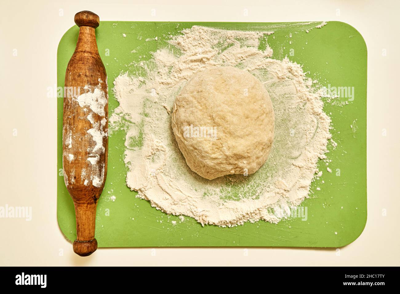 Pezzo di pasta cruda di farina di grano e vecchio mattarello di legno con asse di plastica verde su tavola bianca in cucina vista ravvicinata dall'alto Foto Stock