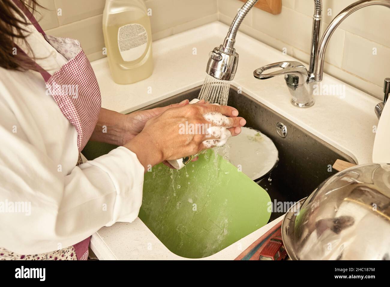 Donna in grembiule e mani schiumose lavano i piatti sporchi con acqua corrente in moderno lavandino in cucina luce vista estrema ravvicinata Foto Stock