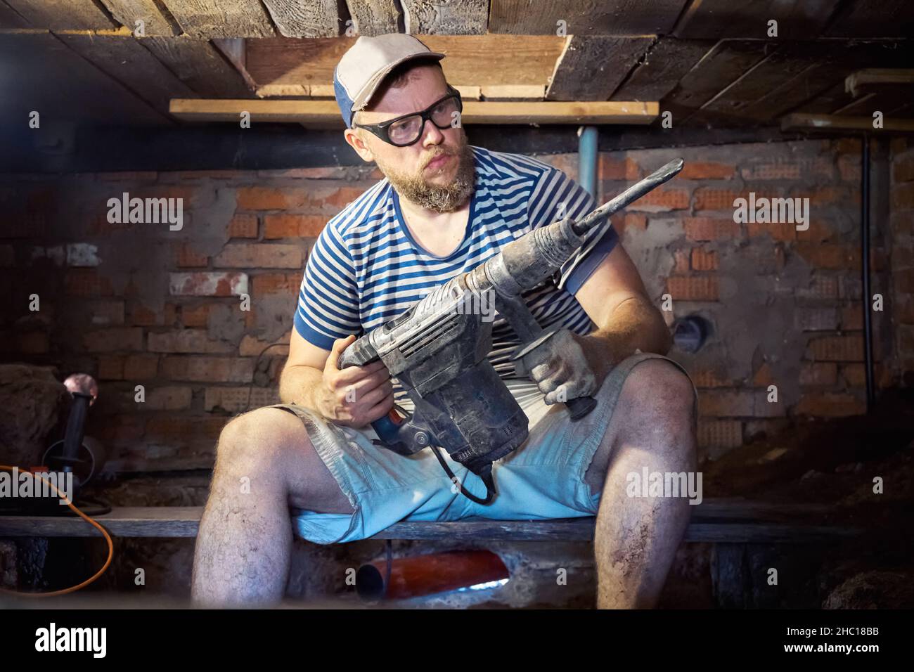 Divertente uomo portico in una T-shirt a righe e occhiali di sicurezza con perforatore per impieghi pesanti ha divertimento durante i lavori di ristrutturazione nel seminterrato della casa. Alto Foto Stock