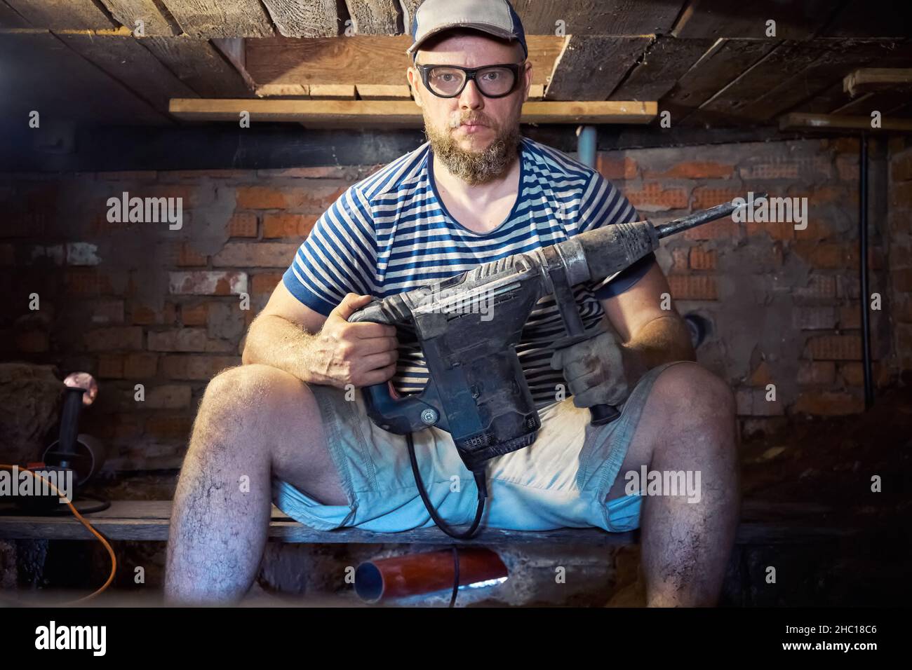 Divertente uomo portico in una T-shirt a righe e occhiali di sicurezza con perforatore per impieghi pesanti ha divertimento durante i lavori di ristrutturazione nel seminterrato della casa. Alto Foto Stock