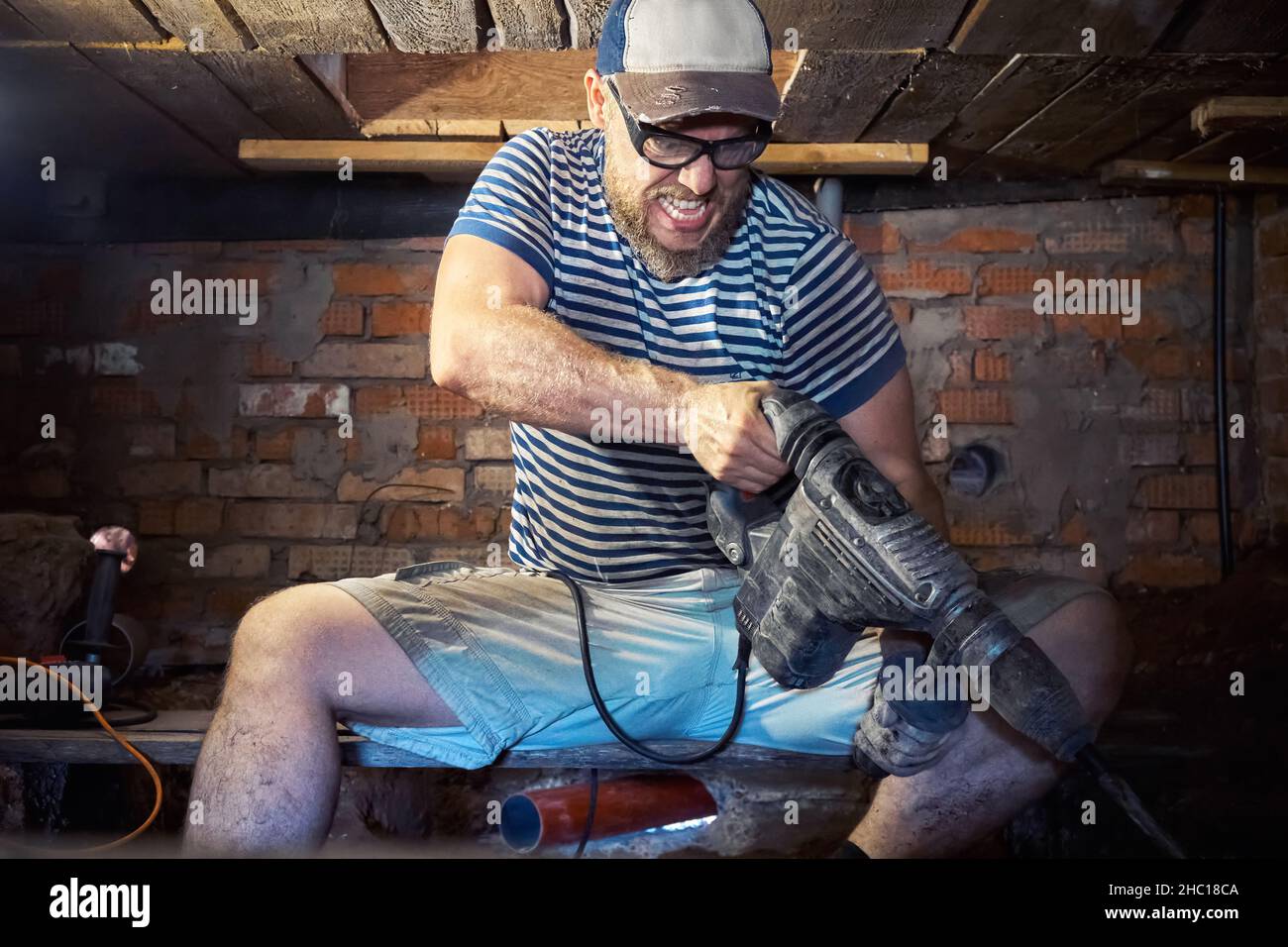 Divertente uomo portico in una T-shirt a righe e occhiali di sicurezza con perforatore per impieghi pesanti ha divertimento durante i lavori di ristrutturazione nel seminterrato della casa. Alto Foto Stock