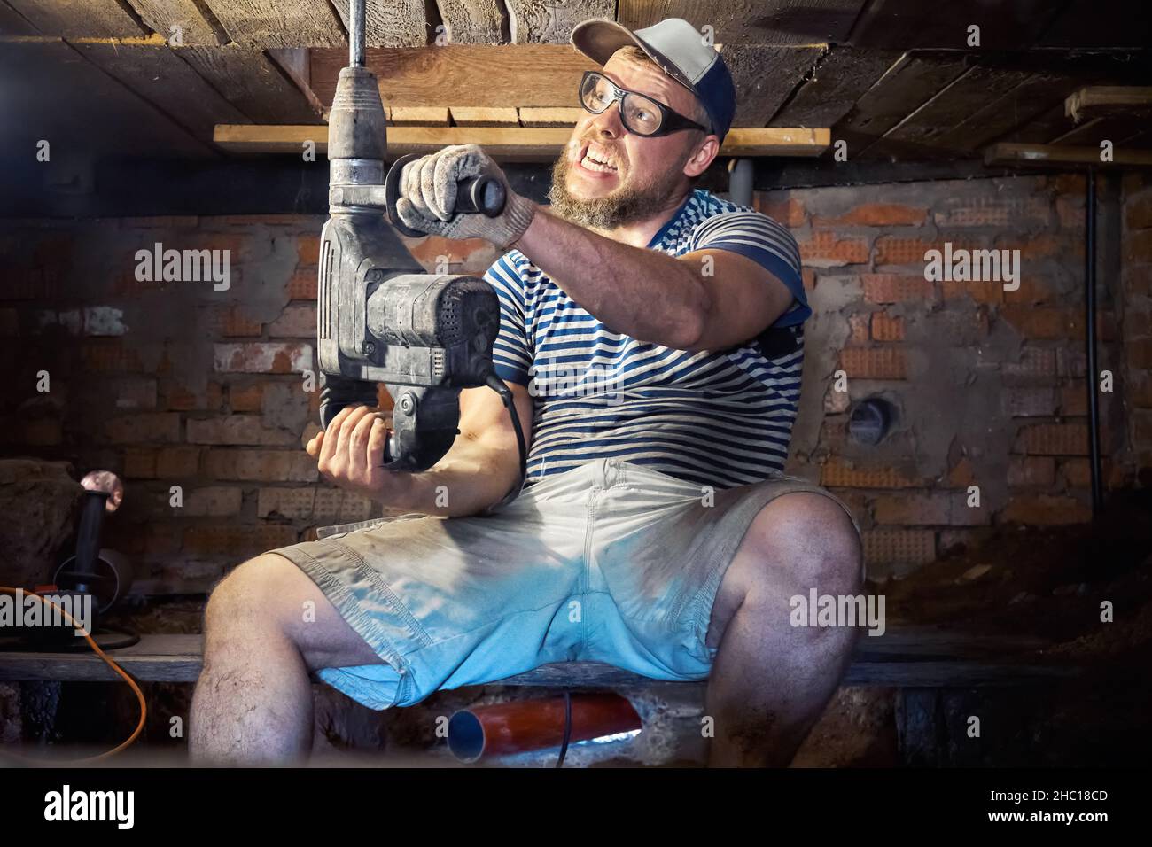 Divertente uomo portico in una T-shirt a righe e occhiali di sicurezza con perforatore per impieghi pesanti ha divertimento durante i lavori di ristrutturazione nel seminterrato della casa. Alto Foto Stock