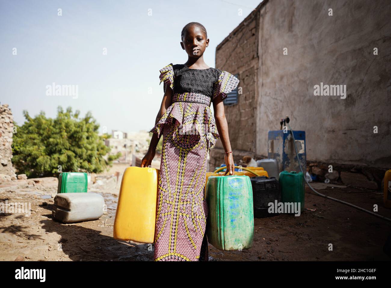Ragazza africana piuttosto nera con due grandi canister d'acqua lasciando il luogo d'acqua pubblica in una mattina calda e soleggiata; concetto di scarsità d'acqua Dometicwater Foto Stock