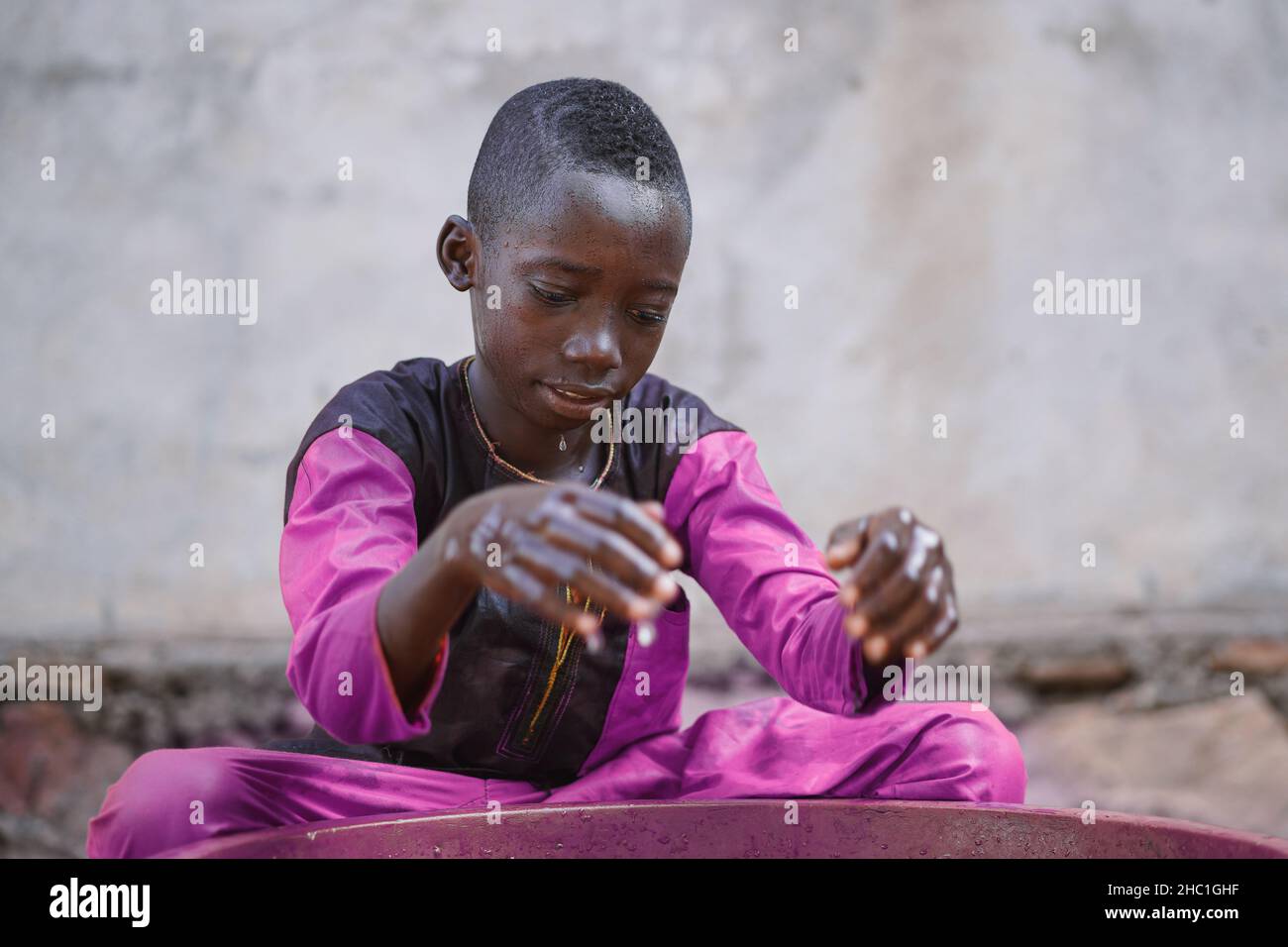 Carino nero africano ragazzo seduto a gambe incrociate di fronte a una vasca di plastica fissando ipnotizzato alla sua riflessione sulla superficie dell'acqua, mentre goccia Foto Stock