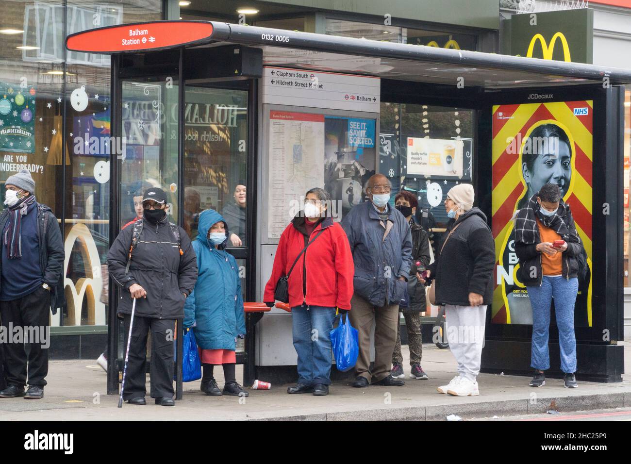 Londra, Regno Unito, 23 dicembre 2021: Le persone si fermano presso una fermata dell'autobus a Balham, già indossando maschere facciali che sono ora obbligatorie per i passeggeri sui trasporti pubblici. Negli ultimi due giorni di shopping prima di Natale i lotti della gente sono fuori e circa l'acquisto dei generi alimentari e dei regali. In Inghilterra non sono state introdotte nuove norme covidi nonostante un numero molto elevato di casi omicron, ma sono attese poco dopo Natale. Anna Watson/Alamy Live News Foto Stock