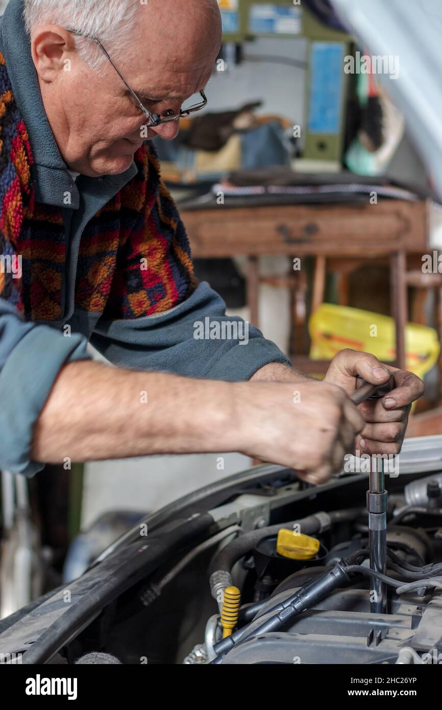 Ritratto di un meccanico senior che cambia le candele sulla sua vecchia auto in garage. Fissare le candele con un cacciavite speciale lungo. Auto vecchie. Foto Stock