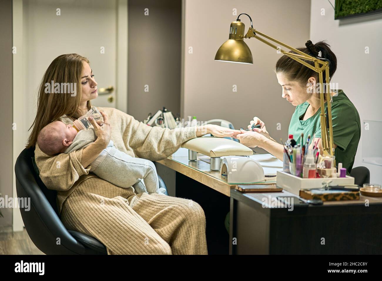 La madre nutre il suo piccolo bambino mentre ottiene un manicure nel salone. Master in guanti blu e verde uniforme facendo manicure sotto la lampada d'oro. Biondo e brunetta. Foto Stock