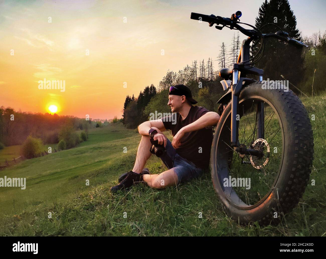 Un uomo è seduto sull'erba vicino ad una bicicletta con ruote spesse. Sullo sfondo del tramonto. Foto Stock