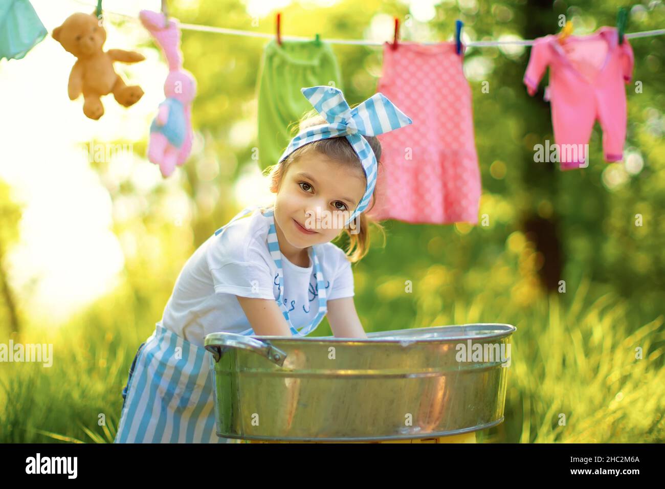 Carina bambina lavando i suoi giocattoli in pelvi in grembiule a scacchi e fascia all'aperto Foto Stock