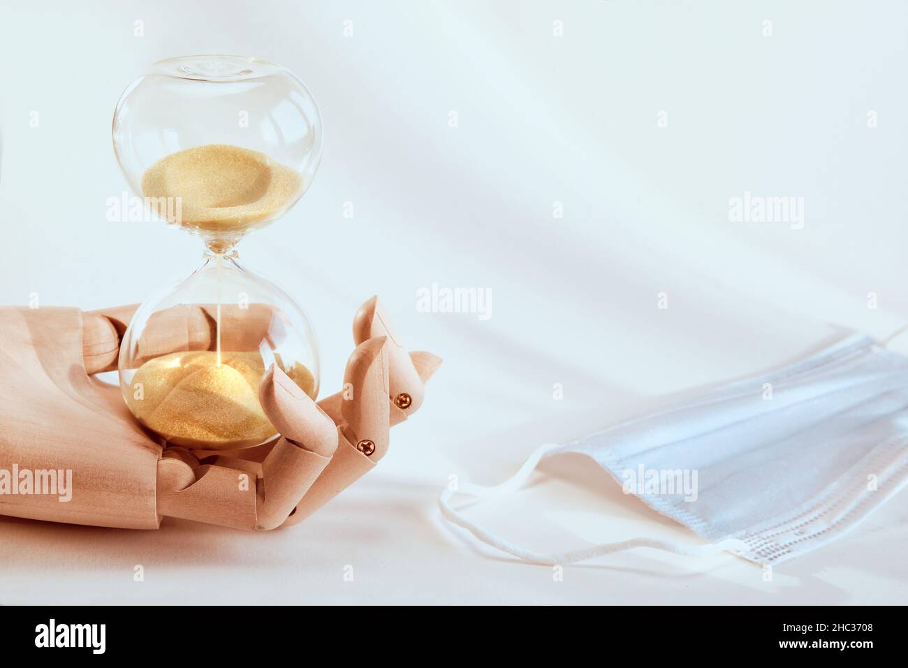 Clessidra in legno modello mano, maschera medica viso. Il concetto di tempo di pandemie di Covid, la sindrome post-covid lunga. Luce naturale con ombre, copia Foto Stock