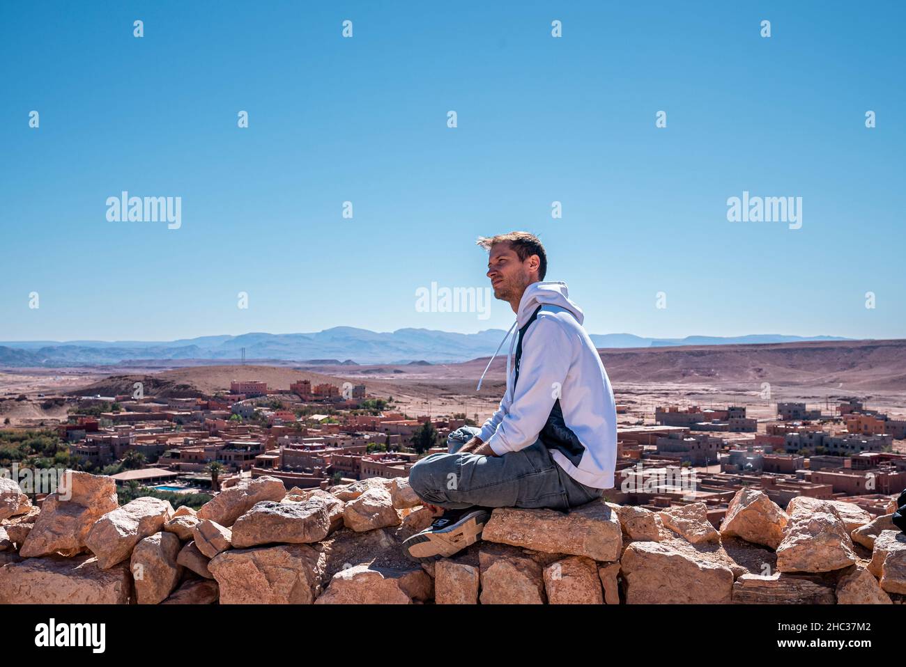 Giovane uomo seduto su pietra contro la vista panoramica della terra deserta e del cielo Foto Stock