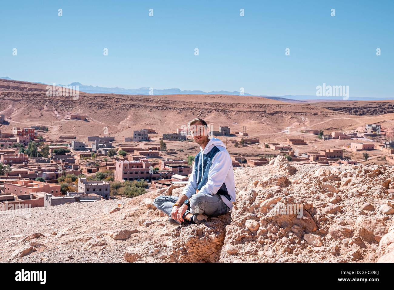 Giovane uomo seduto su roccia contro la vista panoramica della terra deserta e cielo nuvoloso Foto Stock