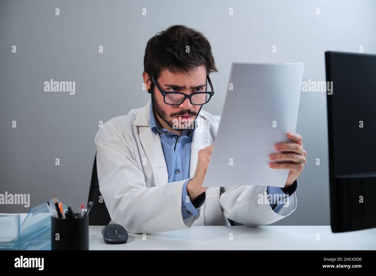Giovane medico che guarda il rapporto medico seduto alla scrivania in un ufficio medico. Foto Stock