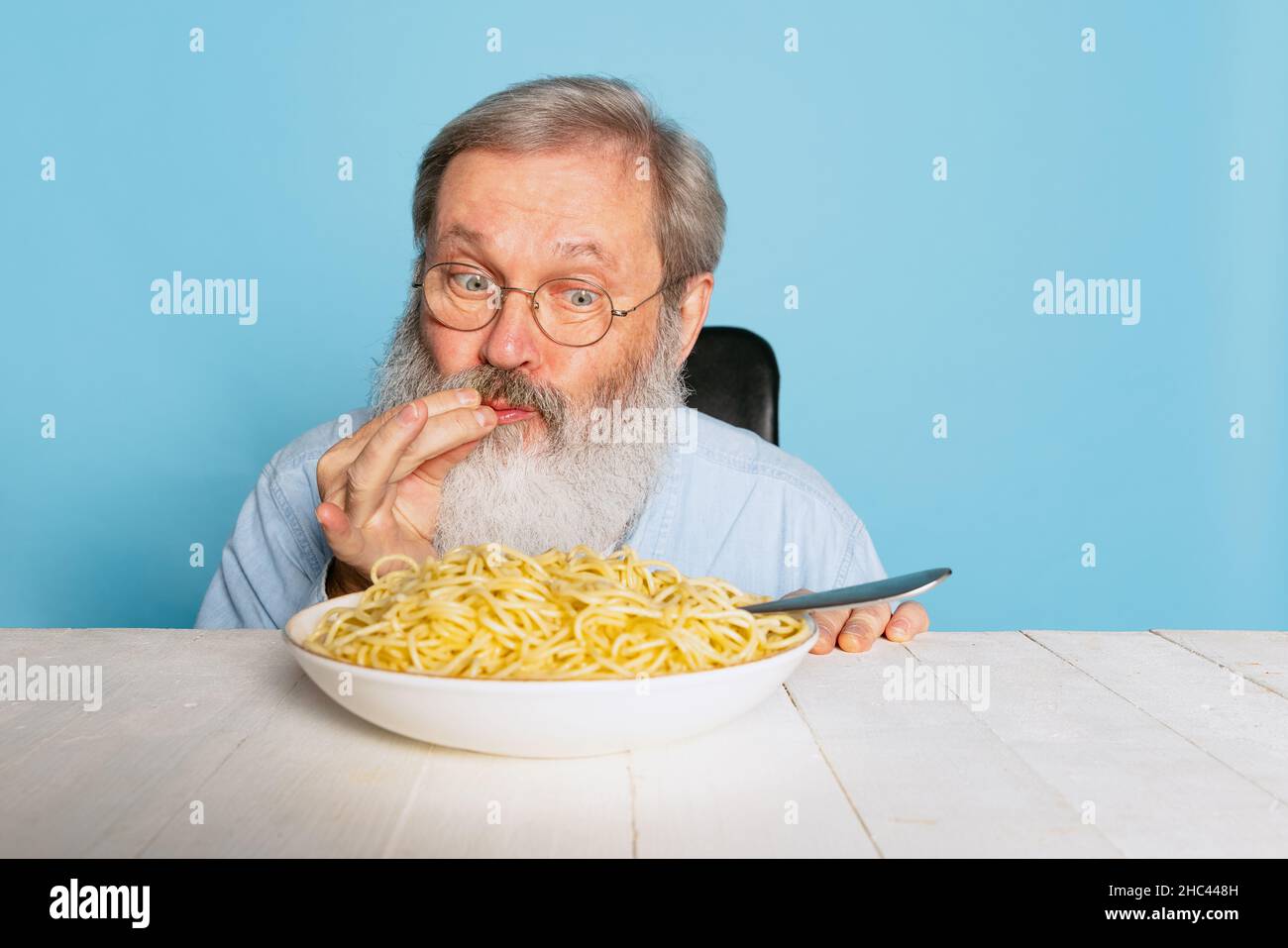 Emotivo uomo peloso senior grigio-bearded mangiare una grande porzione di spaghetti, pasta isolata su sfondo blu studio. Foto Stock