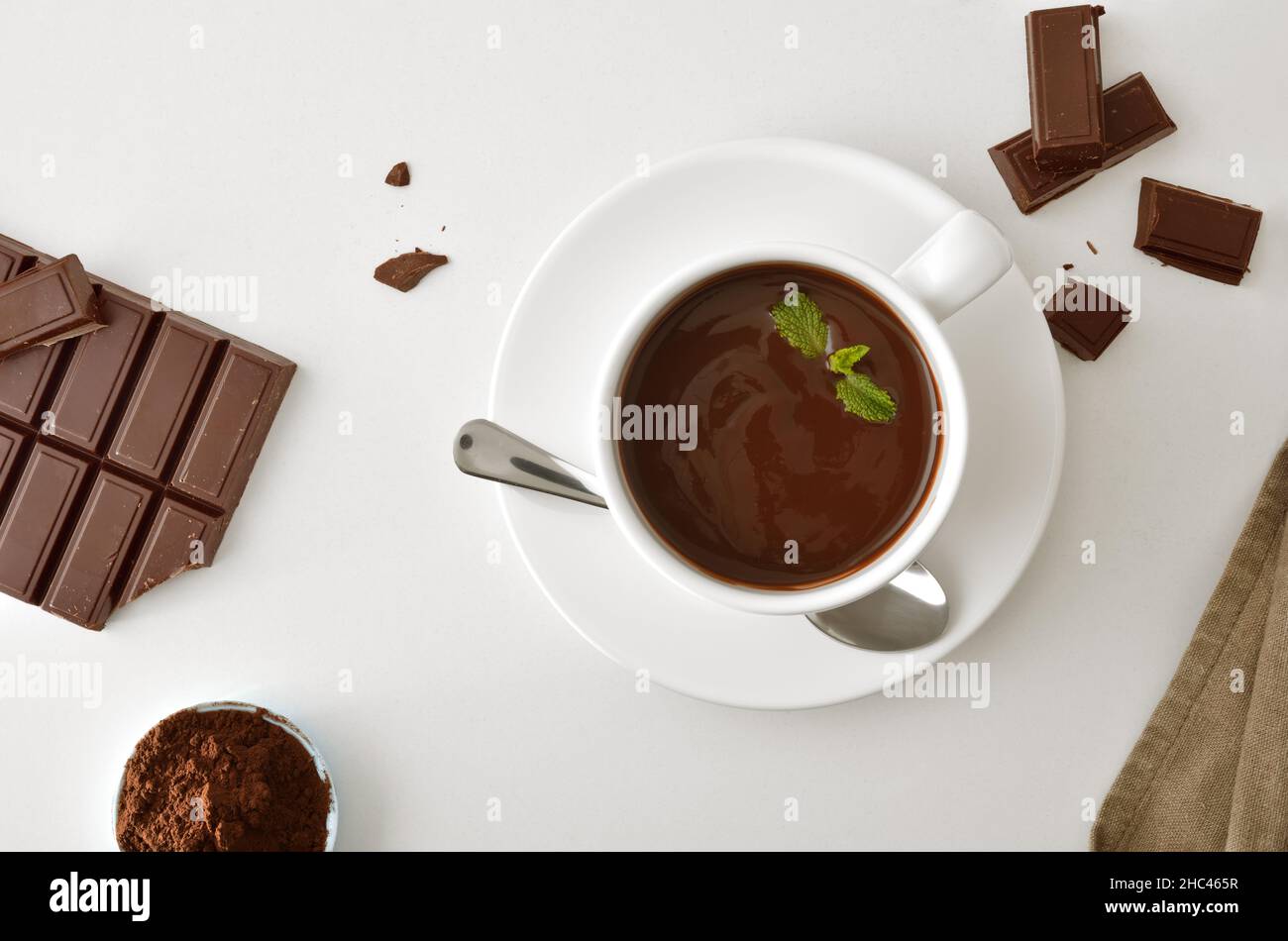 Cioccolata calda in tazza di ceramica bianca, cioccolato dolciario e pezzi e polvere in ciotola su tavola bianca. Vista dall'alto. Composizione orizzontale. Foto Stock