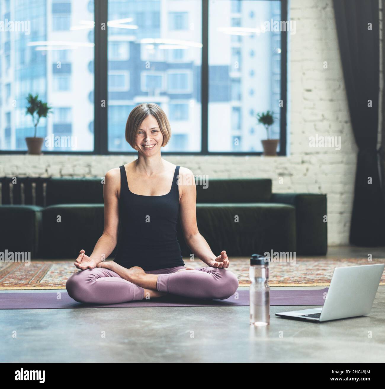 Donna seduta in posa di loto e meditando. È calma, rilassata e felice fuori dalla finestra vivace metropoli. Yoga Donna praticare yoga meditazione esercizio sulla mat nel suo salotto a casa. Primo piano Foto Stock