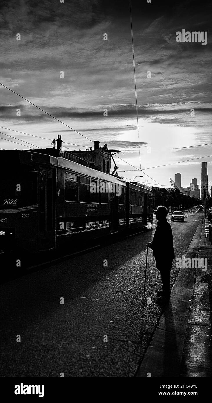 Ripresa verticale in scala di grigi di un vecchio uomo in piedi in strada come un tram passa. Foto Stock