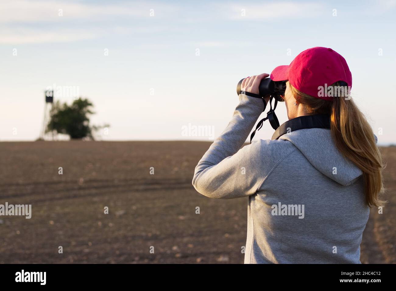 Giovane donna con binocolo che guarda la torre di caccia nel campo. Cacciatore femminile in cerca di una buona posizione per la caccia. Foto Stock