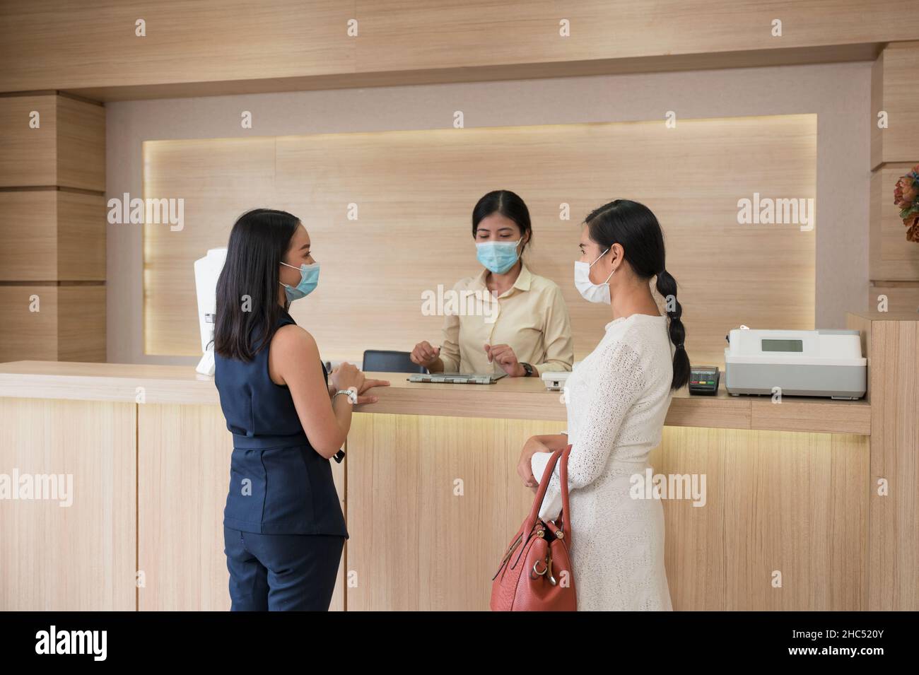 Paziente asiatico donna con maschera facciale con sorella in piedi contatto con il receptionist al banco nella clinica Foto Stock