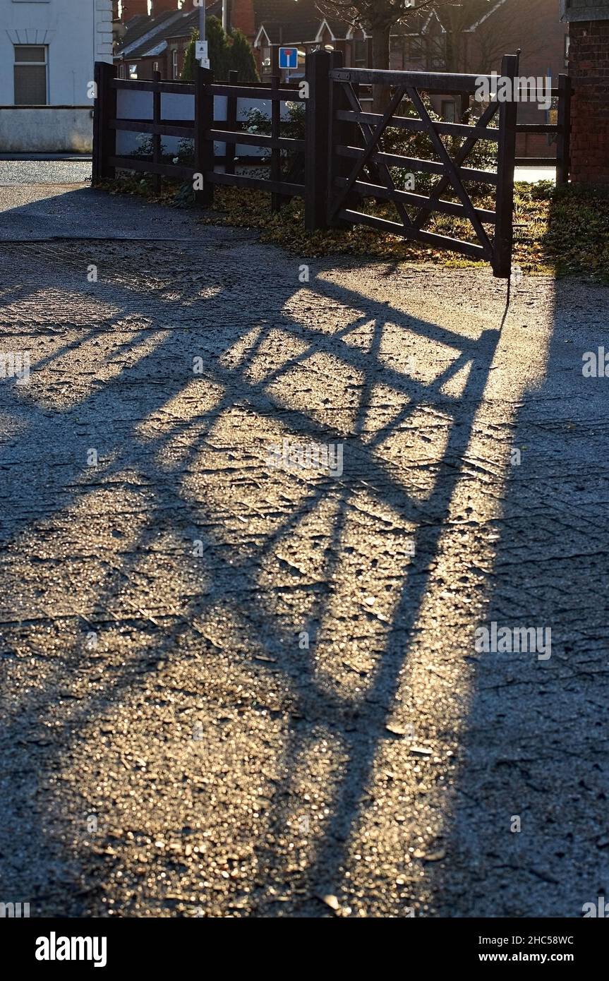 Un cancello di legno aperto che getta lunghe ombre su un terreno di cemento al sole del mattino presto Foto Stock