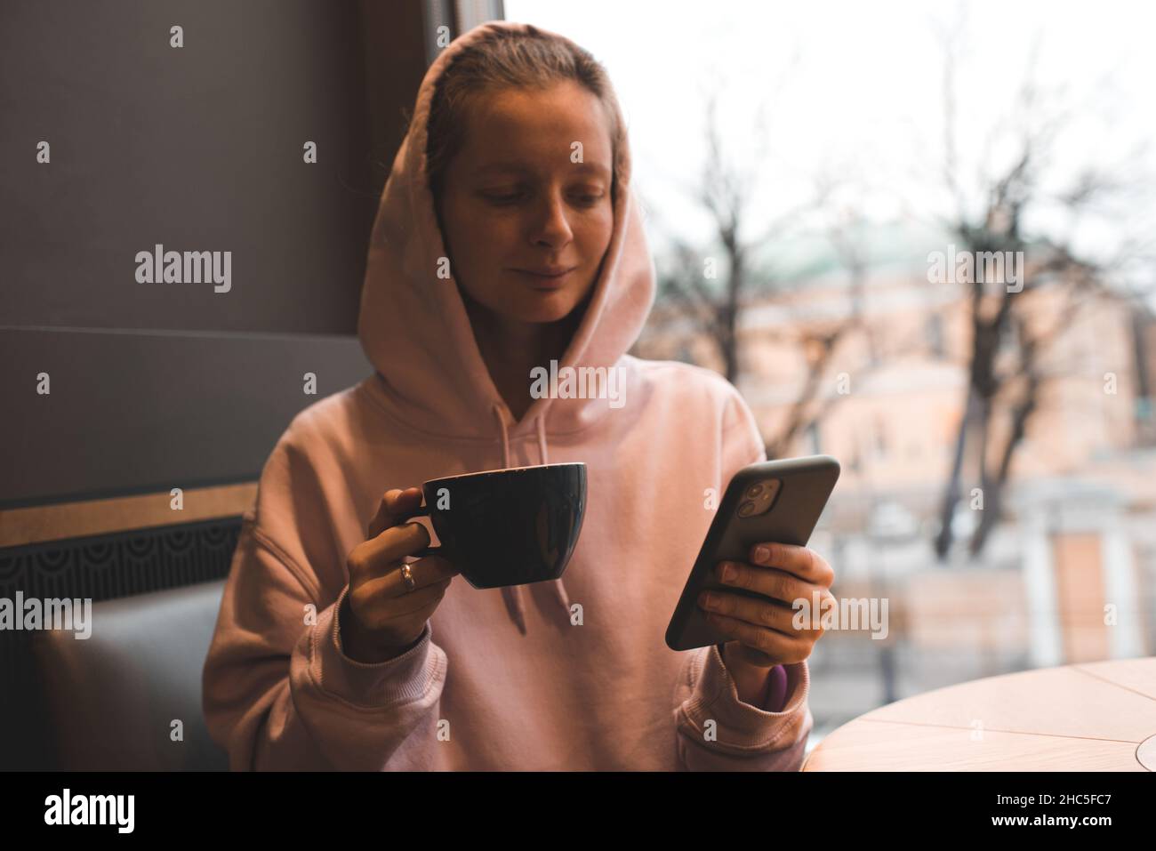 Sorridente giovane ragazza elegante indossare cappuccio bere caffè tenere il telefono in caffè da vicino. Foto Stock