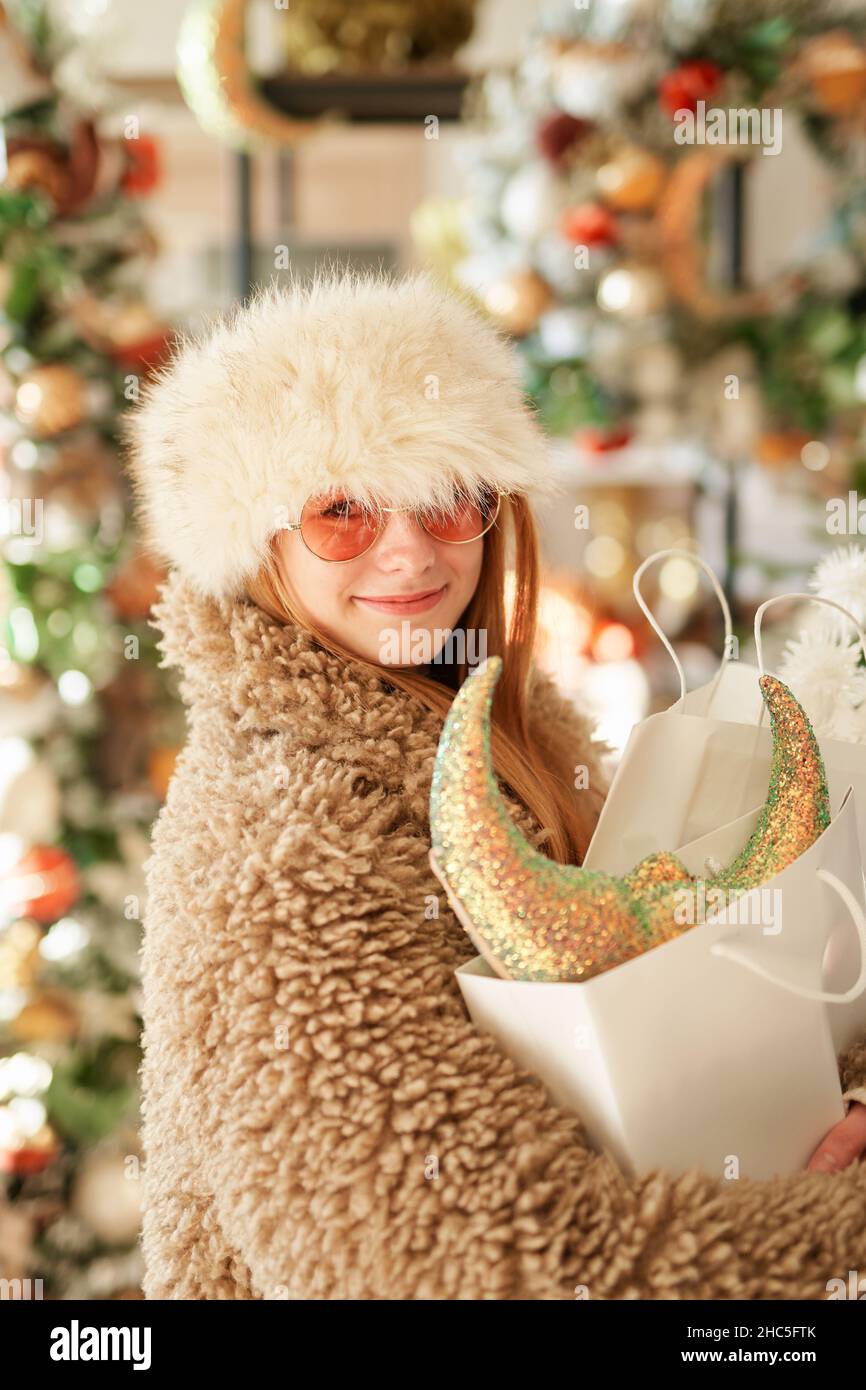 Shopping di Capodanno o tema di consumismo. Ragazza caucasica carina in abiti caldi e occhiali da sole con borse shopping pieno di decorazione di Natale in negozio o supermercato. Immagine di alta qualità Foto Stock