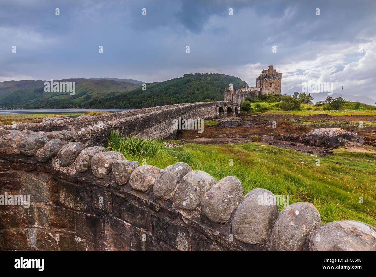 Muro di pietra e ponte di pietra su un accesso all'acqua. Edificio storico Castello di Eilean Donan in estate Vista della fortezza a basse acque con nuvole scure e Foto Stock