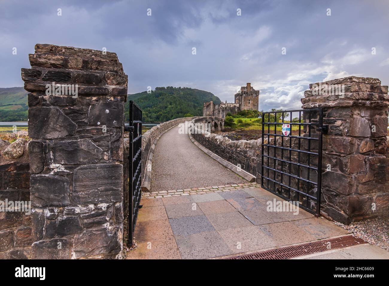Storico muro di roccia con ingresso a un ponte di pietra. Aperto cancello in acciaio nero del ponte pedonale al Castello di Eilean Donan. Colline con alberi e erba verde Foto Stock