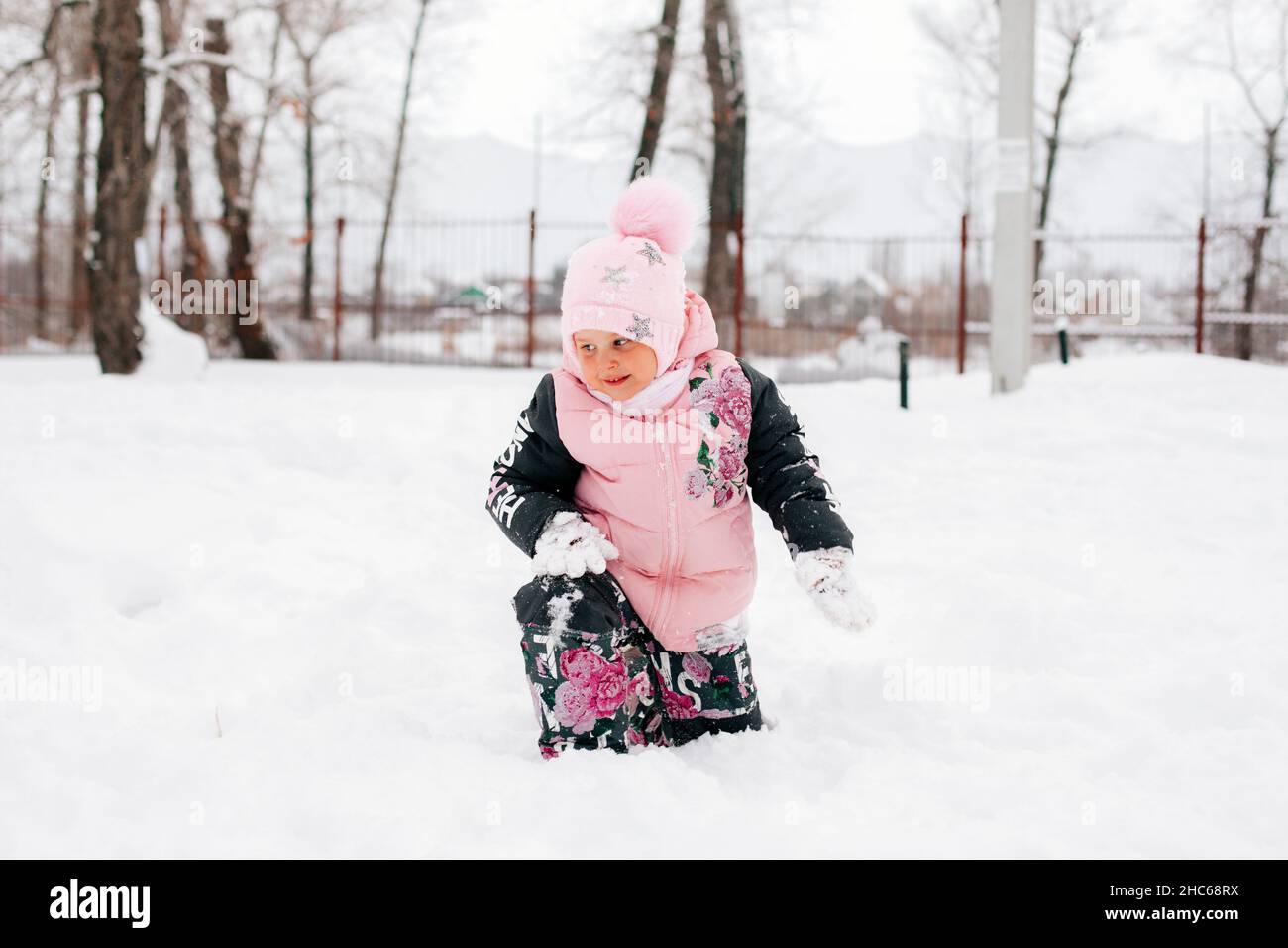 il bambino guarda la neve in piedi da terra e sorride indossando abiti rosa inverno nel parco. Sfondo sorprendente pieno di bianco e neve Foto Stock