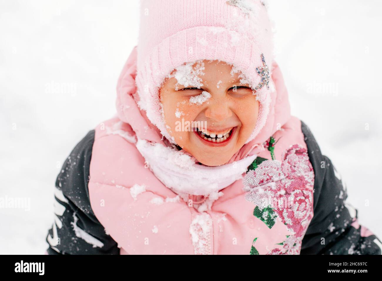 Primo piano bambino sorridente con i denti con il viso coperto di neve indossando abiti rosa inverno nel parco. Sfondo sorprendente pieno di bianco e neve Foto Stock