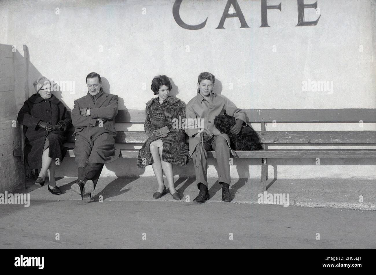 1950s, storico, cappotti, una famiglia britannica, nonna, Madre, padre e figlio adolescente con il loro cane da compagnia, seduto su una lunga panca di legno al di fuori di un caffè sul mare, Inghilterra, Regno Unito. Foto Stock