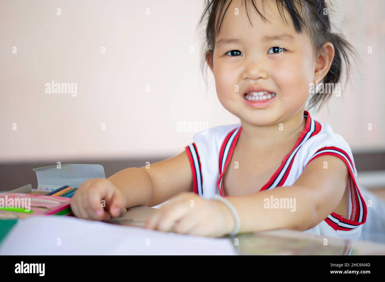 Una giovane bambina asiatica con i capelli neri sorride e mostra dei bei denti, felicemente con matite colorate. Foto Stock