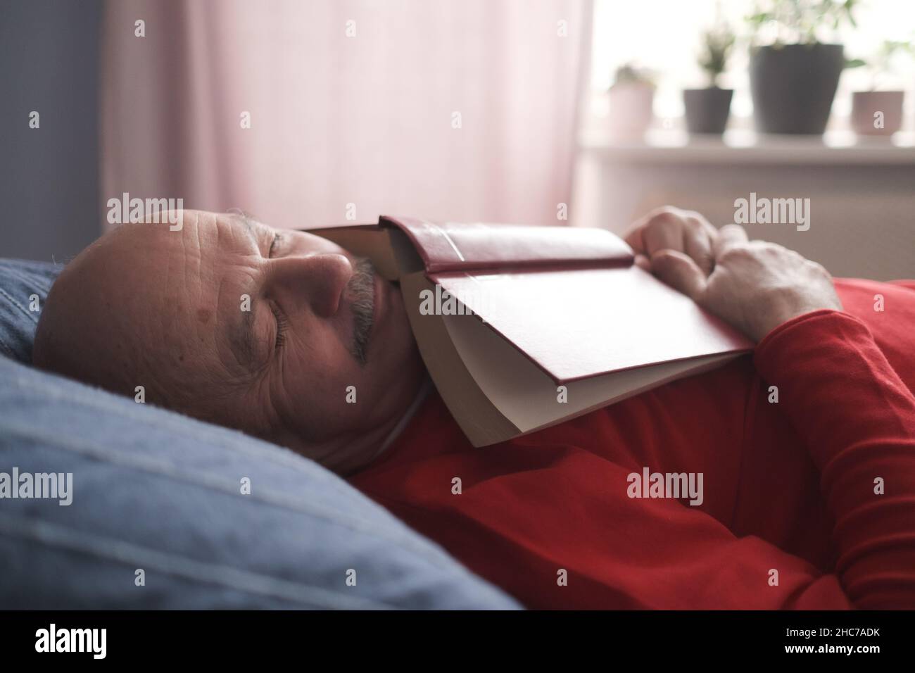 Uomo caucasico anziano che dorme con libro sul divano che peping Foto Stock