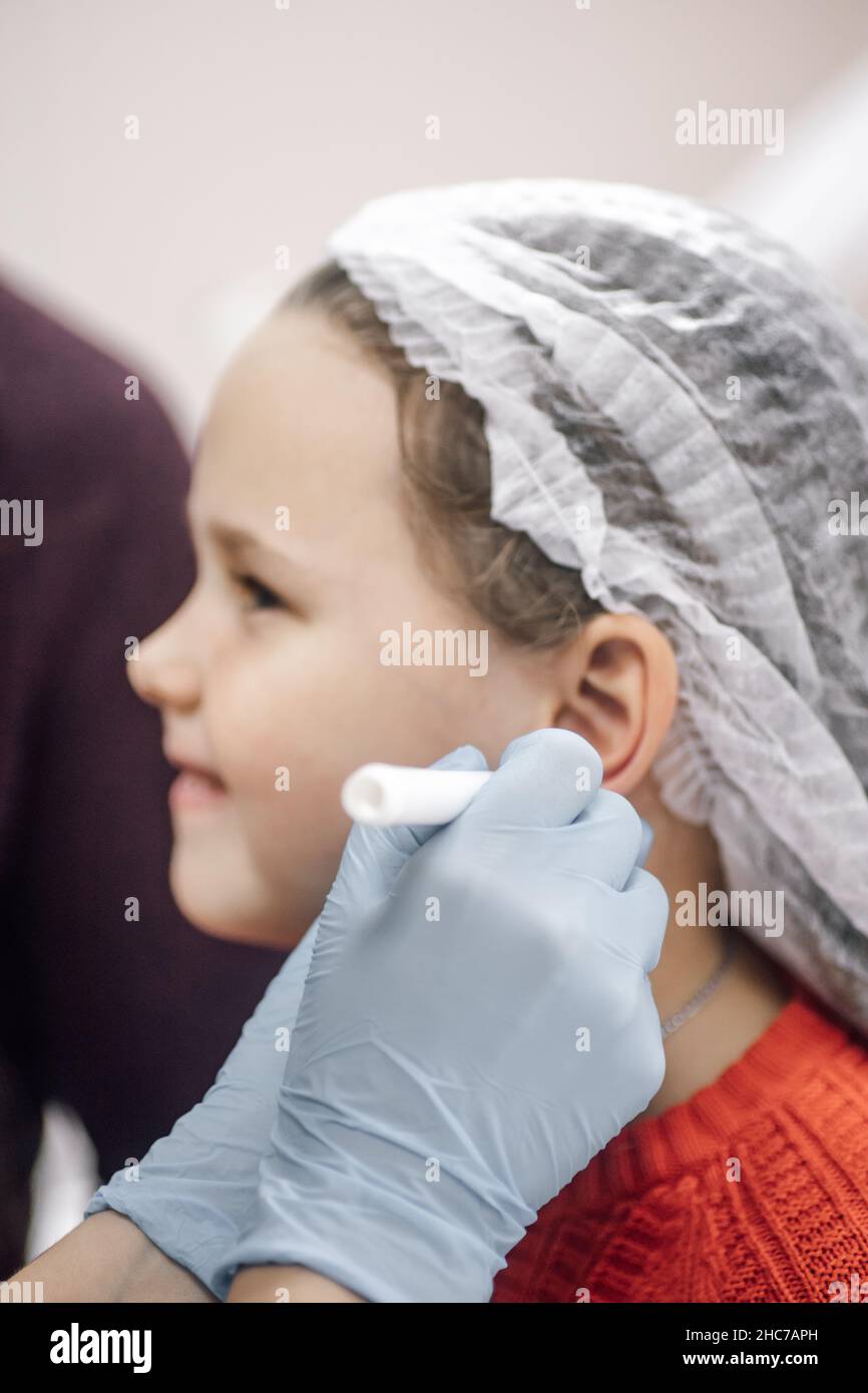 Processo di perforazione dell'orecchio. Il cosmetologo in guanti monouso tira il punto sull'orecchio nel punto di puntura con il marcatore Foto Stock