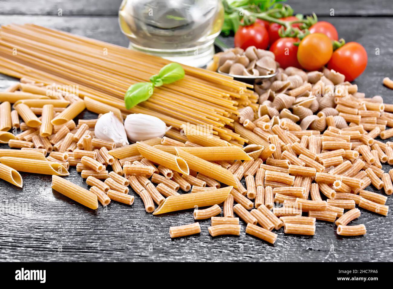 Pasta grano intero e canne di segale rigata, spaghetti, penne; conchiglie, rigatoni, pomodori, aglio, olio vegetale in un decanter su fondo di legno scuro Foto Stock