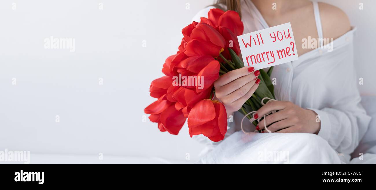Ragazza con fiori e una cartolina mi sposerete su uno sfondo bianco. Bouquet di tulipani rossi nelle mani. Festa concetto San Valentino. Banner. Foto Stock