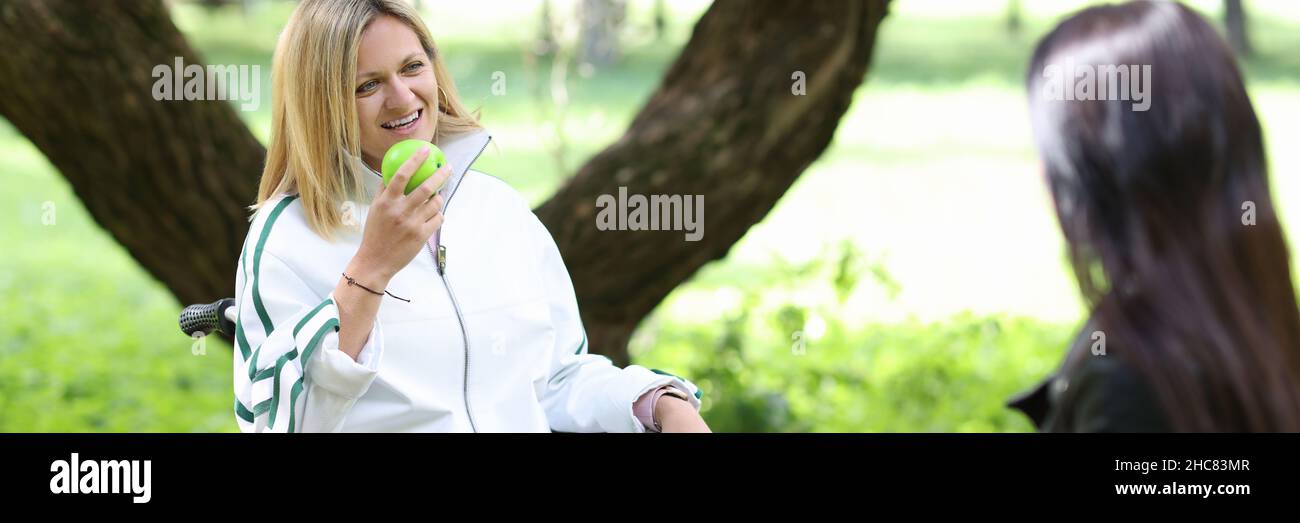 Donna disabile in sedia a rotelle che mangia mela con la sua amica nel parco Foto Stock