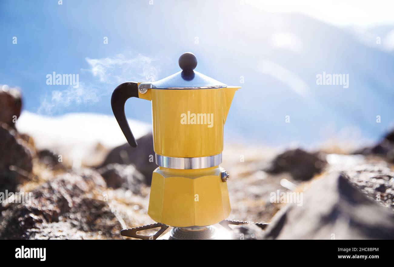 Caffè giallo moka pot con vapore al campeggio neve montagna. Picnic mattutino, persona che cucinano una bevanda calda per colazione nella natura Foto Stock