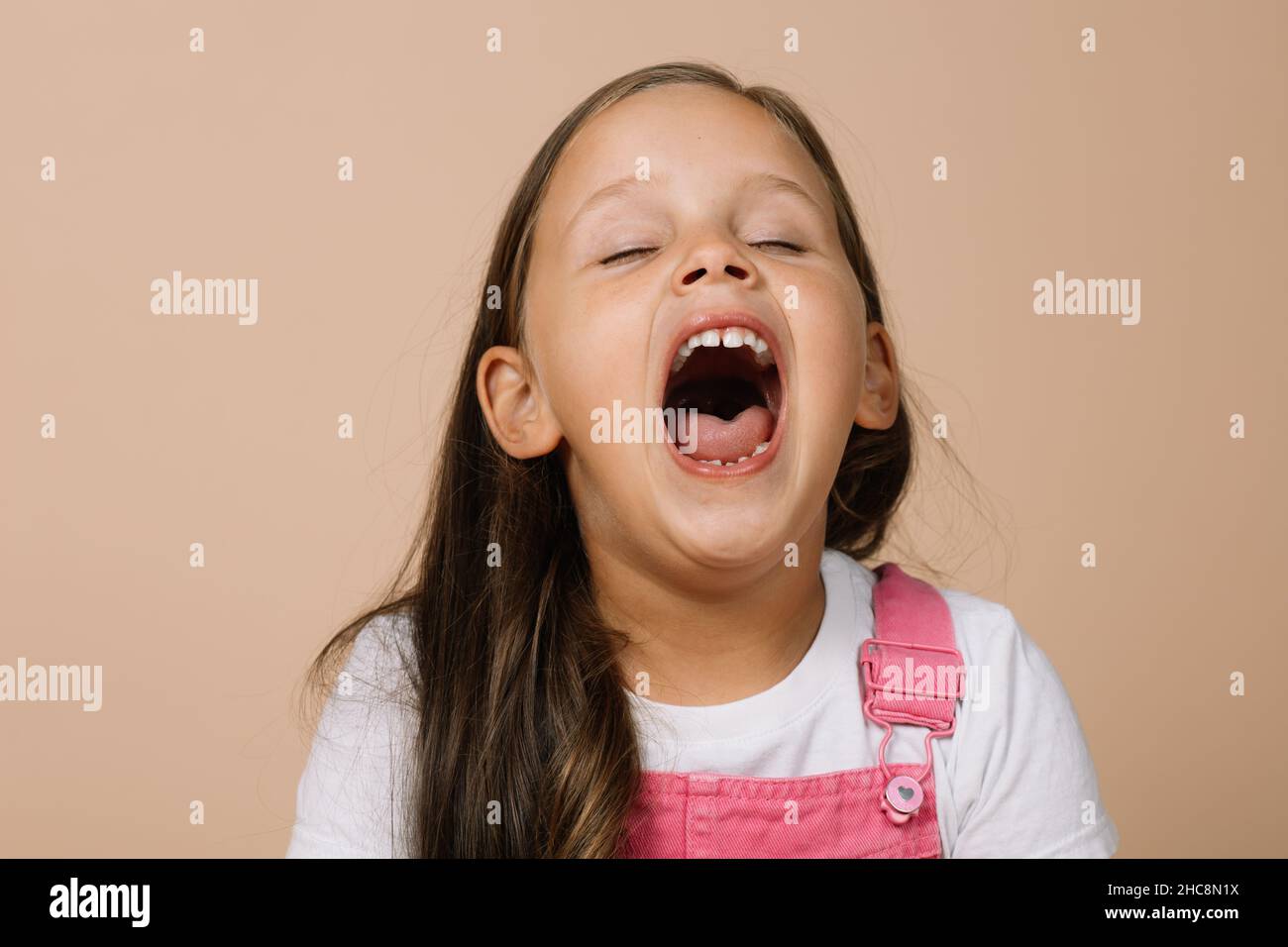 Bambina con bocca completamente aperta e occhi chiusi che sbadigia stanco indossando tuta rosa brillante e maglietta bianca su sfondo beige Foto Stock