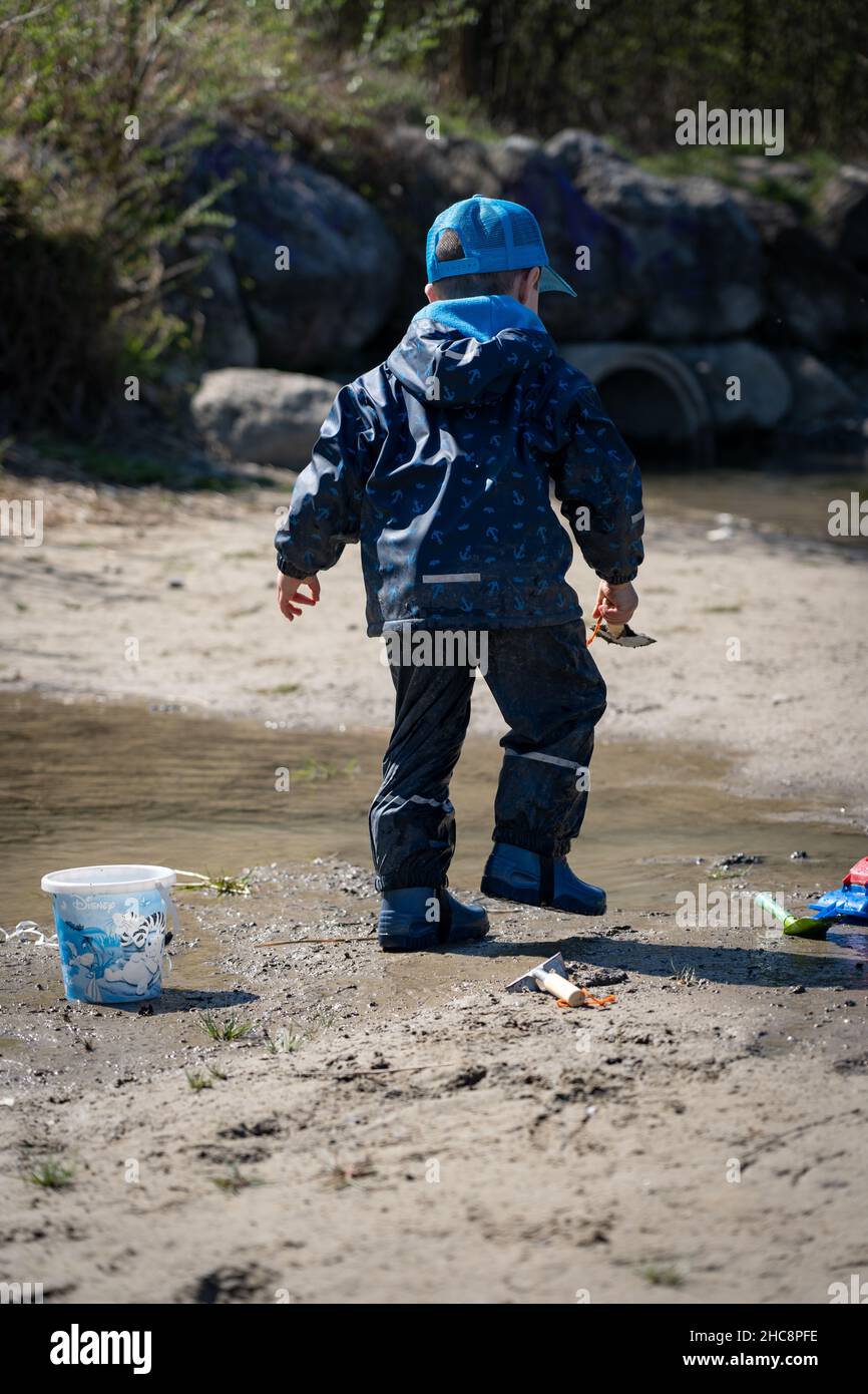 Un ragazzo giovane in un impermeabile blu e stivali da pioggia spruzza felicemente in una pozza d'acqua e fango. Foto Stock